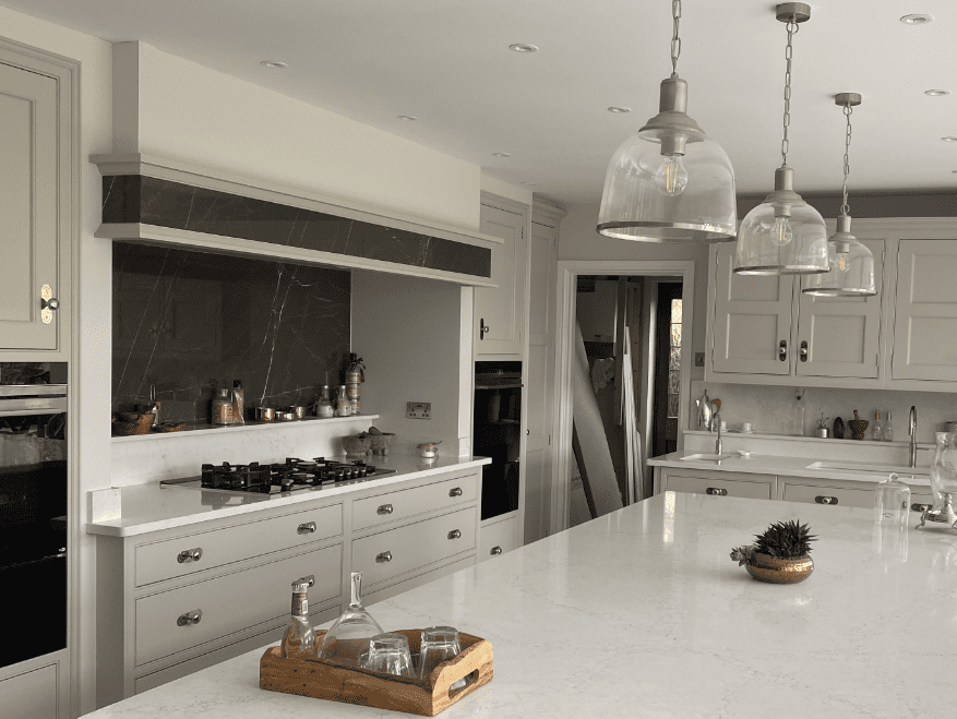 Modern kitchen with a sleek white quartz island, light grey cabinetry, glass pendant lights, and a range cooker with a dark stone splashback.