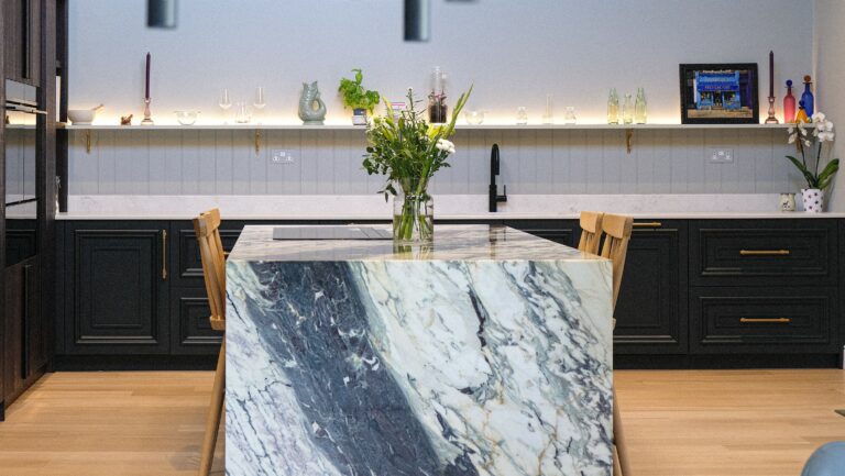 Modern kitchen with a striking marble waterfall island featuring bold veining, paired with black cabinetry, wooden chairs, and a vase of fresh flowers.